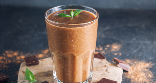 tall glass of creamy chocolate-colored smoothie garnished with fresh mint leaves, sitting on a wooden board with cocoa nibs.